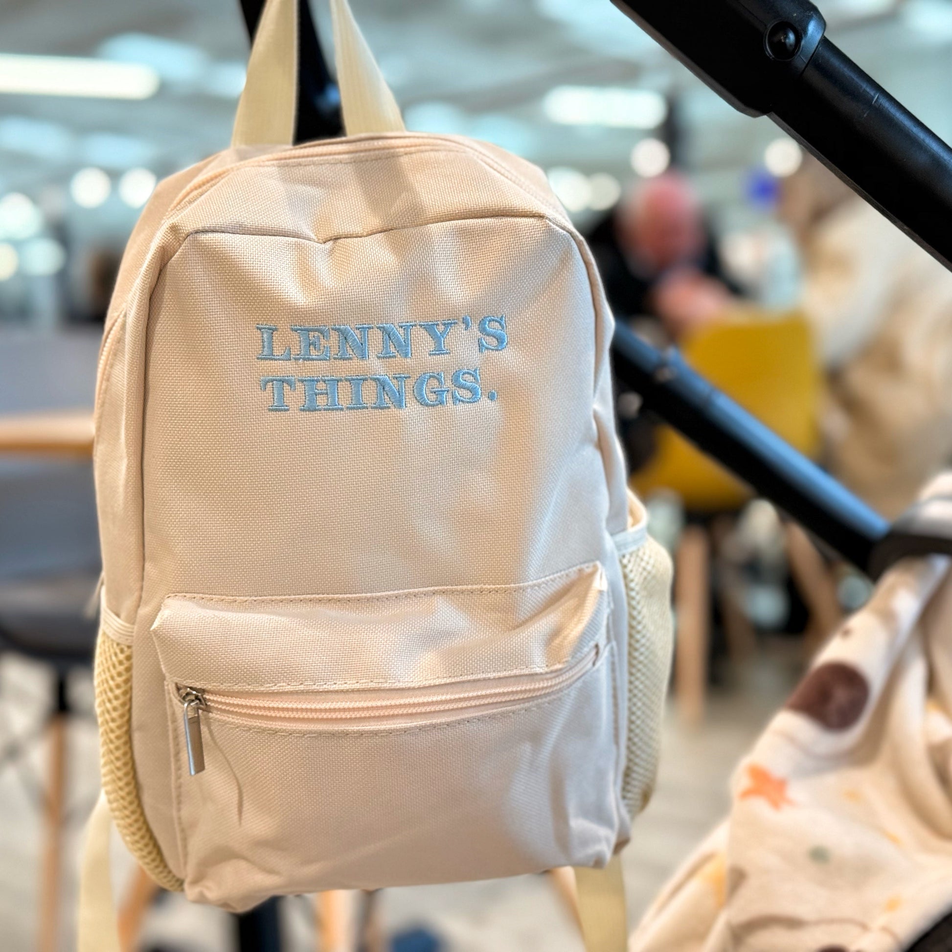 Beige backpack with 'LENNY'S THINGS' text hanging on a stroller in a blurred indoor setting