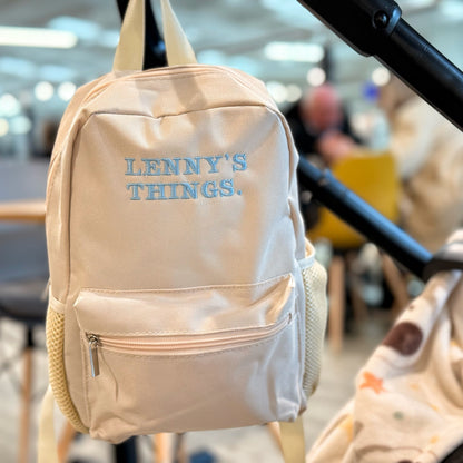 Beige backpack with 'LENNY'S THINGS' text hanging on a stroller in a blurred indoor setting